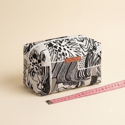 Neceser tipo cubo con estampado abstracto en blanco y negro, cremallera blanca y etiqueta «handmade», junto a una cinta métrica rosa sobre fondo crema